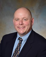 headshot photo of Tim Franz wearing a dark suit jacket, blue checkered shirt and patterned necktie